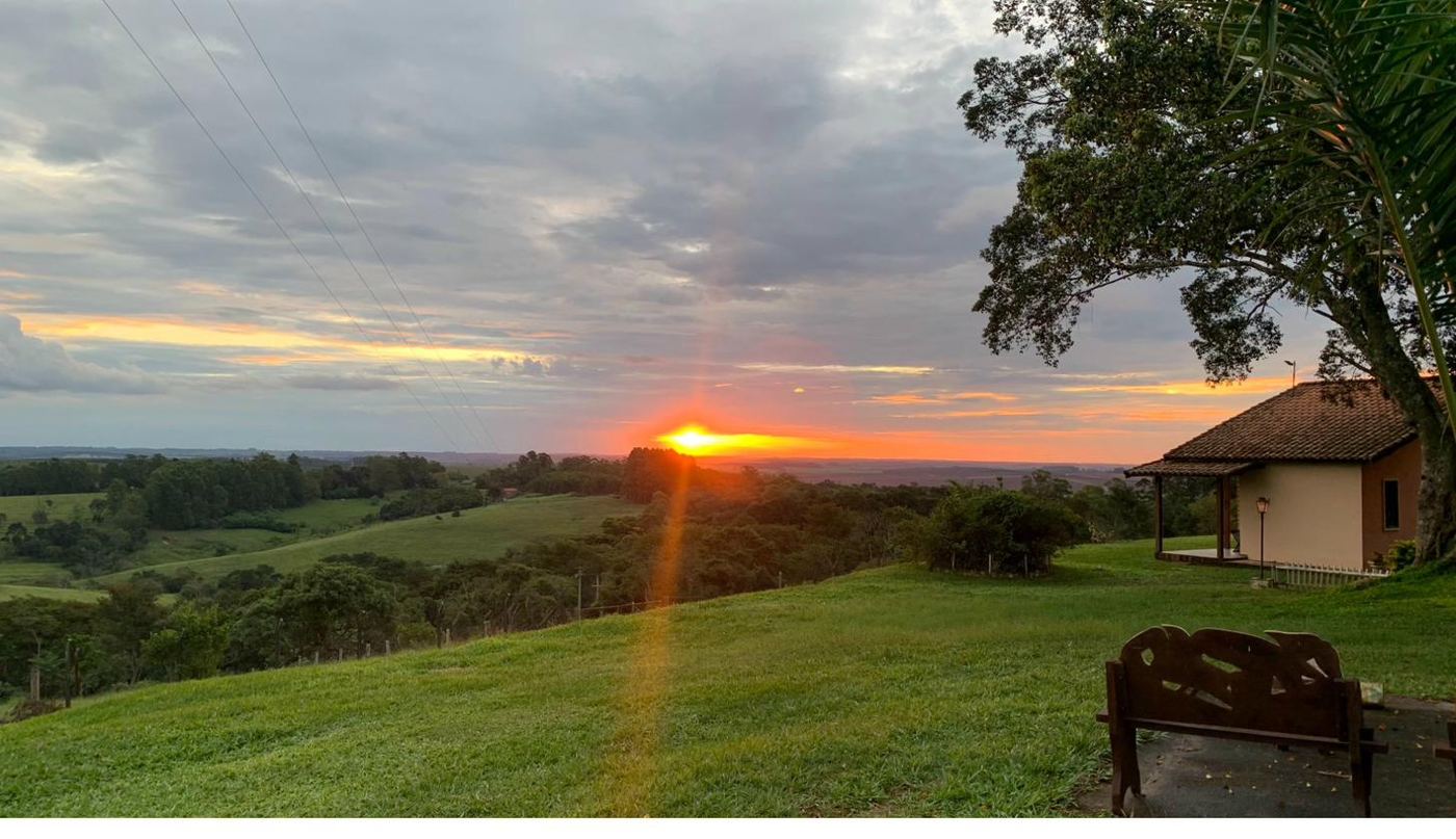 Pôr do sol no Recanto das Seriemas com vista panorâmica do vale, árvore e banco no gramado — pousada na Serra do Itaqueri