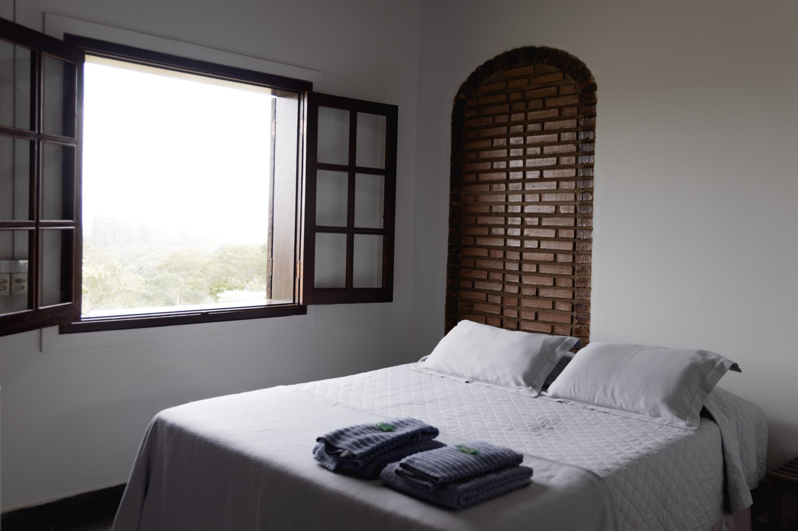 Suíte com cama de casal, nicho de tijolo aparente e janela de madeira aberta para o vale da Serra do Itaqueri — Recanto das Seriemas