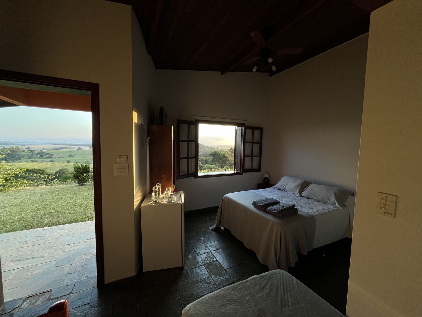 Interior de suíte com cama de casal, lençóis claros, teto de madeira e janela aberta enquadrando o vale da Serra do Itaqueri — Recanto das Seriemas